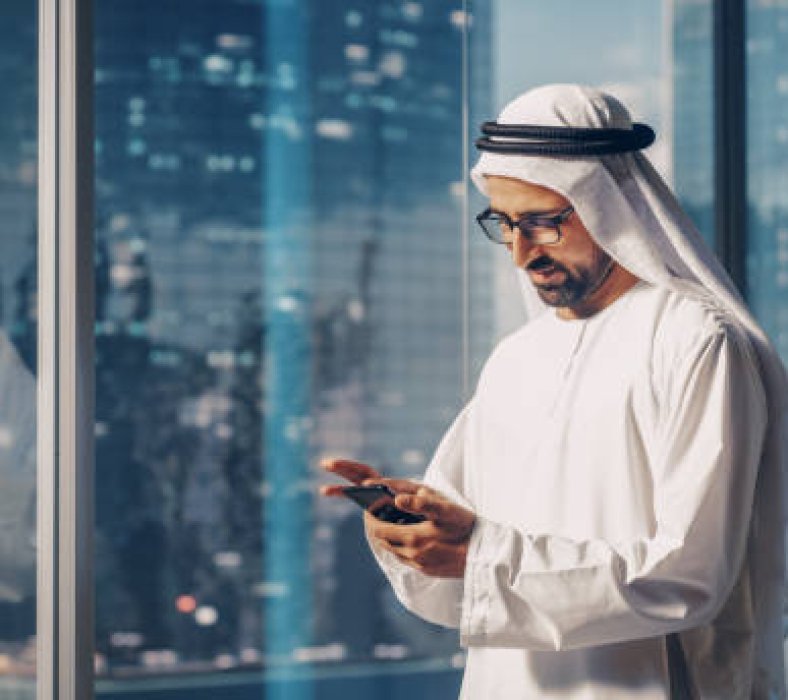 Successful Muslim Businessman in Traditional White Outfit Standing in His Modern Office, Using Smartphone Next to Window with Skyscrapers. Successful Saudi, Emirati, Arab Businessman Concept.